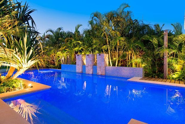 A vibrant outdoor swimming pool surrounded by dense tropical planting. The pool features vivid blue water, tiled edges, a raised wall with vertical stone-clad water features, and a timber deck area, photographed in bright daylight.