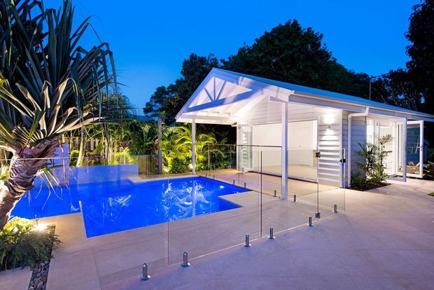 A brightly illuminated modern poolside pavilion at dusk, featuring a white pitched-roof structure with open glazing, horizontal wall cladding, timber decking, and a vivid blue swimming pool surrounded by tropical planting and pale paved surfaces.