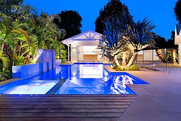 An expansive illuminated swimming pool courtyard at dusk, featuring geometric blue-tiled water features, a timber deck in the foreground, large pale-paved surrounds, tropical planting, and a glazed pavilion or pool house beyond the water. The scene has a polished resort-like appearance with strong blue architectural lighting and reflective water surfaces.