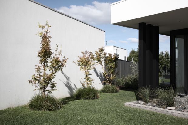 A minimalist modern courtyard or garden beside a house, featuring a white rendered wall, crisp flat roof overhang, black vertical structural columns, dark-framed glazing, manicured lawn, ornamental grasses, and sparse planted beds with gravel edging. The composition emphasizes clean geometric forms and a restrained black-and-white architectural palette.