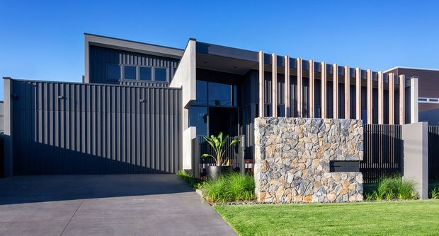 Contemporary two-storey residential facade with a dark rendered and vertically screened exterior, strong geometric forms, timber-toned vertical fins, extensive natural stone feature cladding, and a recessed entry. A broad concrete driveway and manicured lawn frame the street-facing frontage.