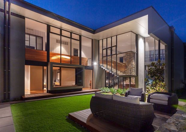 A contemporary multi-level courtyard-facing home photographed at dusk, with a sharply folded flat roof, extensive floor-to-ceiling glazing, dark metal-framed windows, warm timber interiors, and a dramatic open stair. The landscaped courtyard includes a lawn, timber deck, outdoor seating, and built-in planting.