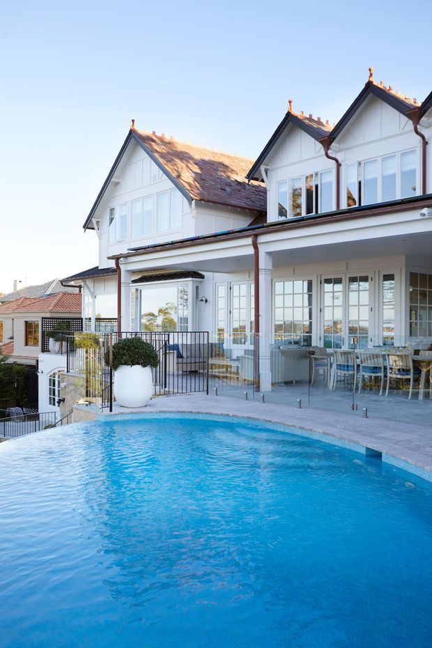 Exterior view of a large white multi-storey house with steep gabled roofs, decorative gable detailing, extensive glazed windows and doors, and a covered outdoor dining area. A swimming pool occupies the foreground, with a paved terrace, glass balustrade, planters, and landscaped levels around the house.