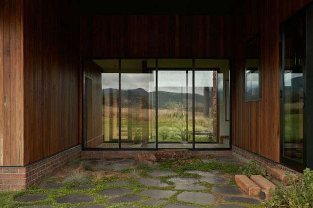 A dark timber-clad entry courtyard or covered outdoor threshold, framed by glazed walls and doors. The space has a low red-brick plinth, irregular stone paving with vegetation growing between slabs, and a view through the glazing toward a rural, mountainous landscape. The design is restrained and strongly connected to the outdoors.
