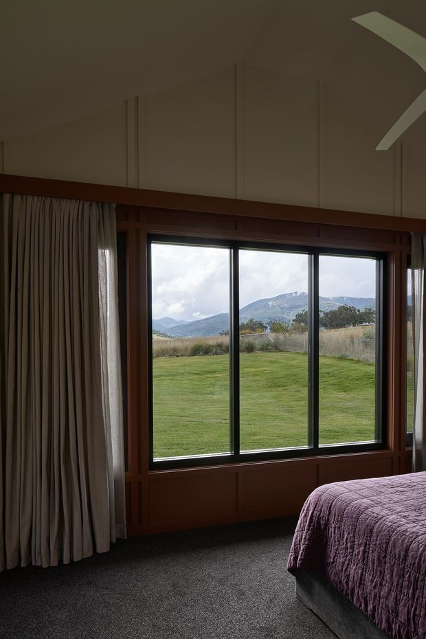 A dim, warmly toned bedroom with a large dark timber-framed picture window overlooking a grassy rural landscape and mountain range. Full-height pleated curtains frame the glazing, while a partially visible bed with a deep purple quilt occupies the foreground. The room has dark carpet, timber wall panelling and a high, subtly detailed ceiling.