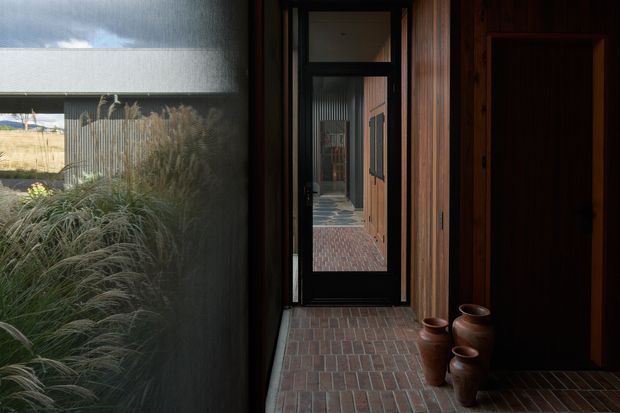 A dark, intimate residential entry hall with a long brick-paved floor, rich vertical timber wall lining and dark timber doors. A glazed internal door creates a strong view through the passage, while a large window on the left looks onto ornamental grasses and a sandy coastal landscape. Terracotta pots line the floor beside the doorway, adding a warm natural accent.
