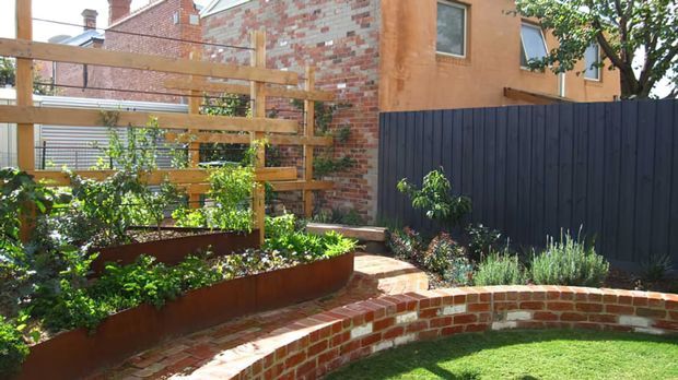 A compact landscaped courtyard garden with raised rusted-metal planting beds, timber trellis screening, curved red-brick retaining walls and steps, lawn, and dense ornamental planting. A dark vertical-board fence and brick residential buildings form the backdrop.