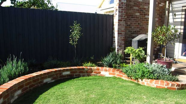 A compact landscaped courtyard garden beside a brick house, featuring a curved low red-brick retaining wall enclosing a neatly trimmed lawn, dense ornamental planting, and a dark vertical privacy fence. The house edge includes exposed brickwork, a covered outdoor area, timber decking, and potted plants.