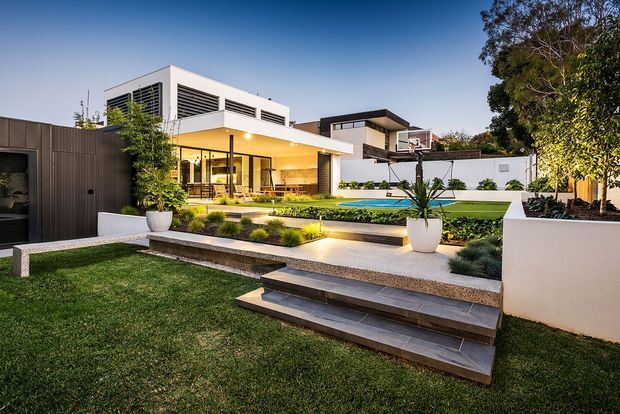 Contemporary multi-level residence viewed from a landscaped backyard, featuring crisp white rendered walls, dark vertical screening, extensive glazing, a broad flat roofline, and a covered outdoor entertaining area. Layered stone-paved terraces and steps lead through manicured lawn and planting toward a narrow swimming pool, with warm architectural lighting creating an inviting evening atmosphere.