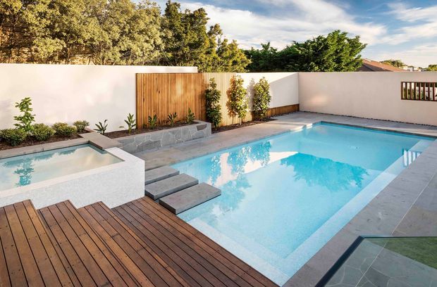 A contemporary landscaped backyard centered on a bright swimming pool with an integrated raised spa, timber decking, broad pale stone paving, stepping slabs, rendered privacy walls, and vertical timber screening. Planting beds and mature trees frame the outdoor entertaining area.