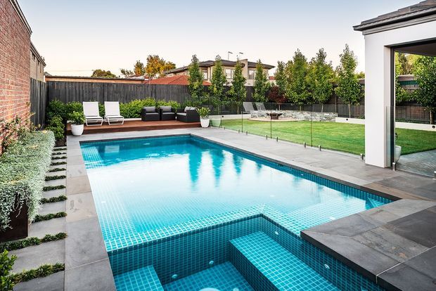 A landscaped residential backyard with a large rectangular swimming pool finished in vivid blue mosaic tile. The pool includes submerged steps and a shallow platform, with pale stone paving around it. A manicured lawn, planted borders, privacy fencing, outdoor loungers, dark built-in seating, and a contemporary white poolside pavilion frame the scene.