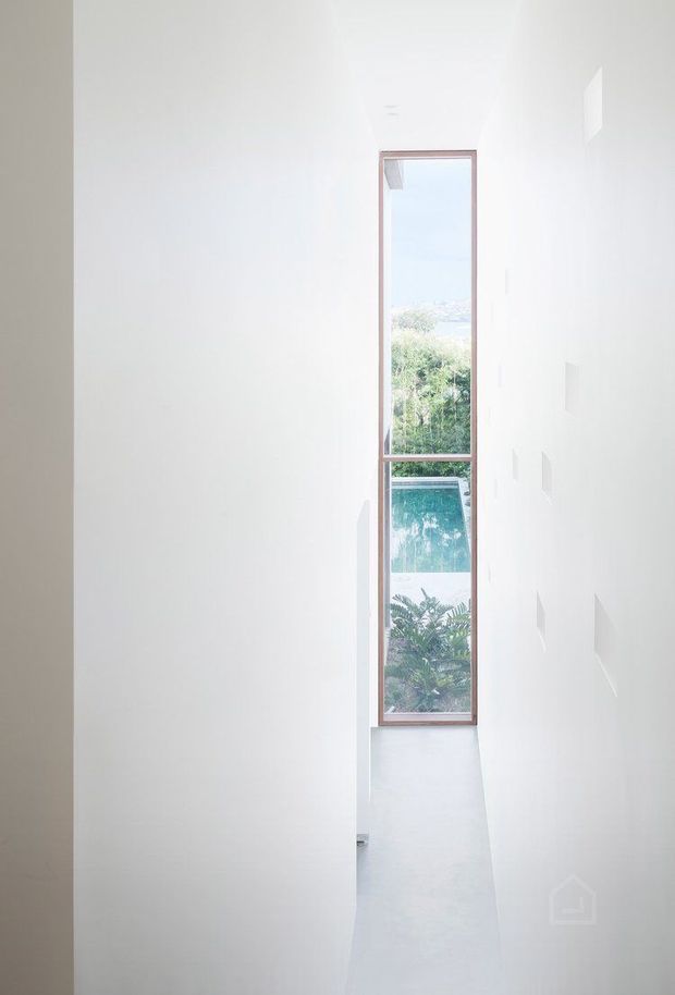A narrow, minimalist white interior passage or stairwell with very tall smooth walls and a slim full-height timber-framed window at the end. The glazing overlooks lush vegetation and a visible swimming pool, bringing daylight into the otherwise stark space. Small recessed wall openings add subtle architectural detail.