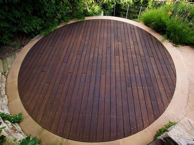 A landscaped outdoor area featuring a circular dark timber deck or platform, bordered by pale paving and surrounded by dense green planting and ornamental grasses.