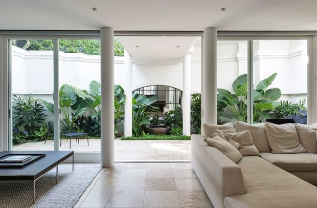 Bright contemporary living room opening onto a lush enclosed courtyard garden. The space features full-height glazed sliding doors, white cylindrical columns, pale stone tile flooring, a large cream sectional sofa, and a low dark coffee table. A covered outdoor passage and arched opening are visible beyond the courtyard.