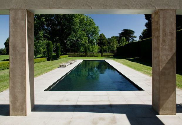 A formal rectangular swimming pool viewed through a broad stone or rendered architectural opening. Pale paving surrounds the dark reflective pool, with neatly clipped hedges, lawns, mature trees, and loungers creating a structured garden setting. Strong sunlight and deep shadows emphasize the framed, symmetrical composition.