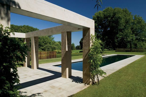 A landscaped outdoor pool terrace framed by a substantial geometric concrete pergola or colonnade. Pale stone or concrete paving surrounds a rectangular swimming pool, with clipped greenery, lawn, mature trees and a timber fence beyond. The composition is strongly linear and architectural under clear blue skies.