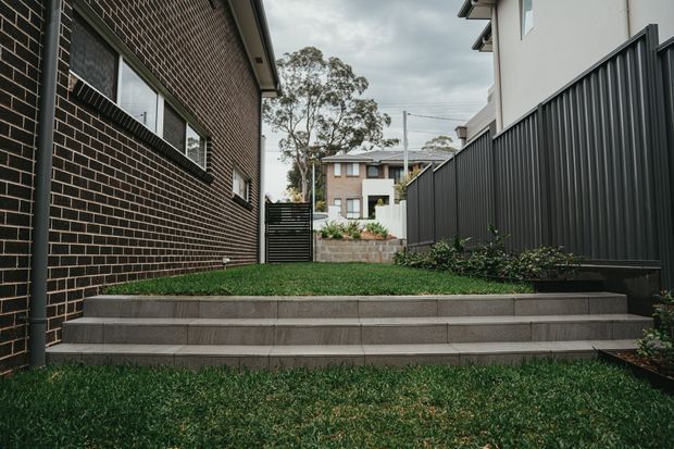 A landscaped side passage between modern residential buildings, featuring broad grey tiled steps, a small lawn terrace, dark brick wall with horizontal windows, and a tall black vertical-panel privacy fence. Low planting borders the fence, with neighboring homes visible beyond.