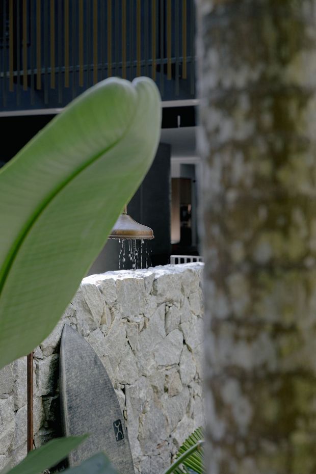A close-up architectural view of a contemporary courtyard or outdoor area, partially framed by tropical foliage and a tree trunk. A rough natural-stone masonry wall supports a small water feature, with a dark modern building facade and vertical timber or metal screening visible behind it. A surfboard leans against the wall, adding a coastal lifestyle element.