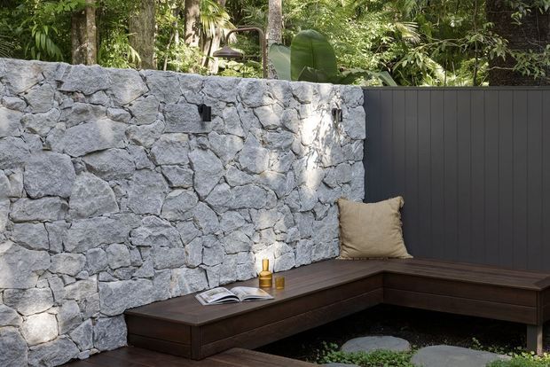 A tranquil outdoor courtyard seating area with a dark timber built-in corner bench, a pale irregular natural-stone wall, and a dark vertical-panel privacy fence. Dense tropical foliage rises behind the enclosure, while a small book, candle, and cushion add understated comfort.
