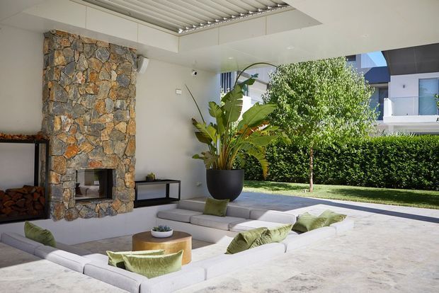 Bright contemporary outdoor lounge beside a covered patio, featuring a large natural-stone fireplace, low modular upholstered seating arranged around a round timber coffee table, a potted tropical plant, and views toward a landscaped garden and neighbouring modern architecture. The space has a partially open roof with a linear skylight or louvred ceiling detail and a polished, resort-like feel.