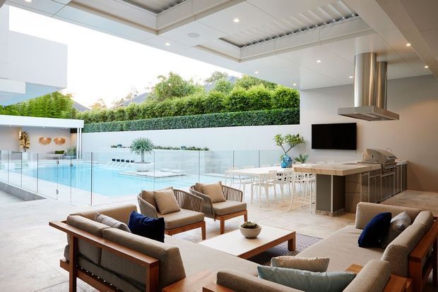 Bright, open-plan contemporary living and dining area with a fully equipped kitchen, timber-framed upholstered seating, a large coffee table, and floor-to-ceiling glazing opening onto a glass-edged swimming pool and landscaped courtyard. The space features a coffered and louvred ceiling, light neutral finishes, extensive glazing, and a statement stainless-steel rangehood over the island.