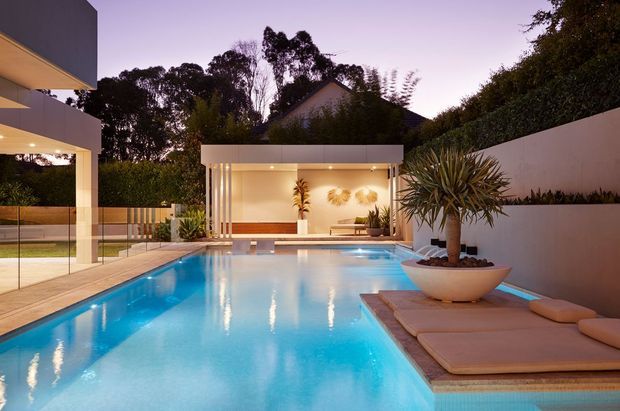 Twilight view of a contemporary residential pool courtyard. A large rectangular swimming pool dominates the foreground, bordered by pale paving and a floating timber-toned sun lounge platform with cushions. The space includes a covered outdoor entertaining pavilion with warm recessed lighting, glass balustrades, raised planters, architectural white walls, and sculptural potted plants.