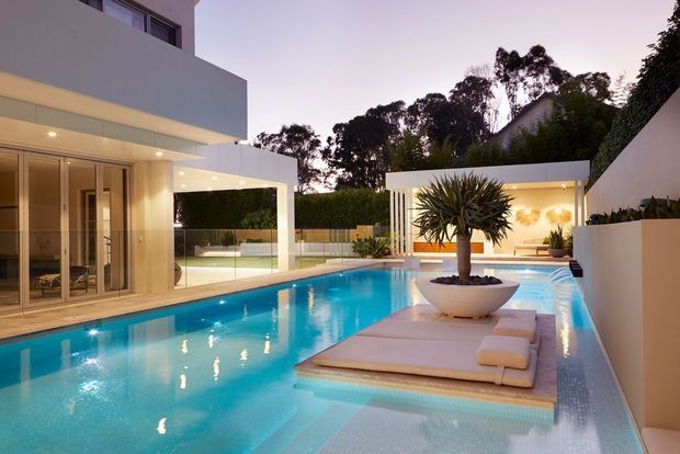 Contemporary luxury residential pool courtyard at dusk, featuring a long illuminated swimming pool, integrated shallow lounging platform, floating deck with a large planter, pale stone or concrete paving, glass balustrades, and crisp white rectilinear architecture with extensive glazing and covered outdoor entertaining areas.