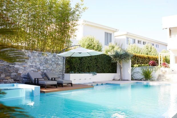 Bright contemporary residential pool area with a turquoise swimming pool, raised spa, timber pool deck, pale stone retaining wall, sculptural planting, hedges, sun loungers, and umbrellas. White multi-level buildings and outdoor landscaping surround the pool, creating a polished resort-like setting.