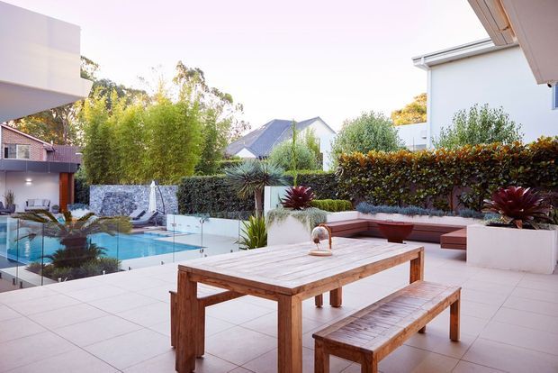 A spacious contemporary outdoor terrace overlooking a landscaped swimming pool courtyard. The tiled patio features a substantial timber dining table with matching bench seating, built-in raised planters, hedging, and modern white rendered buildings surrounding the pool area.