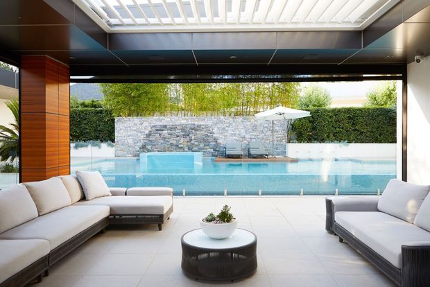 Open-plan indoor-outdoor living area with a contemporary black-framed opening onto a swimming pool. The space features pale upholstered sofas, a round dark coffee table, large light floor tiles, warm timber wall panelling, and a bright overhead glazed skylight or louvred roof structure. Beyond the pool is a stacked-stone privacy wall, landscaping, and outdoor seating.