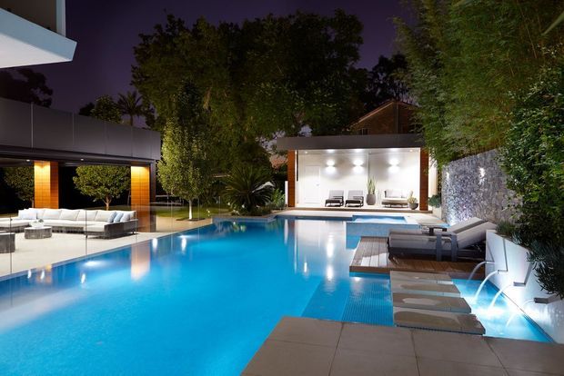 A luxurious landscaped outdoor pool area at night, featuring a large illuminated swimming pool, modern poolside lounge furniture, a timber-look deck, lush planting, and contemporary pavilion-like structures with extensive glazing and warm lighting.