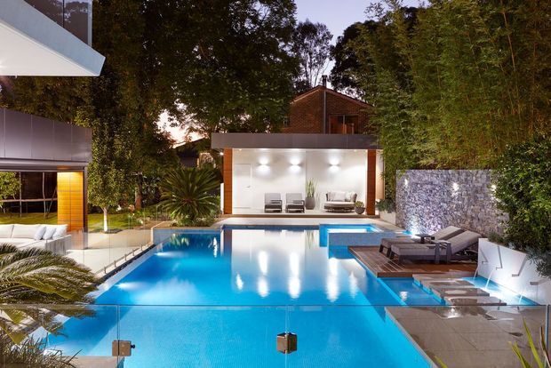A contemporary landscaped residential pool area at dusk, featuring a large illuminated swimming pool, timber pool deck, submerged loungers, a covered outdoor seating pavilion, and a textured stone retaining wall. Dense planting surrounds the space, with portions of the modern house visible at the left and behind the pavilion.