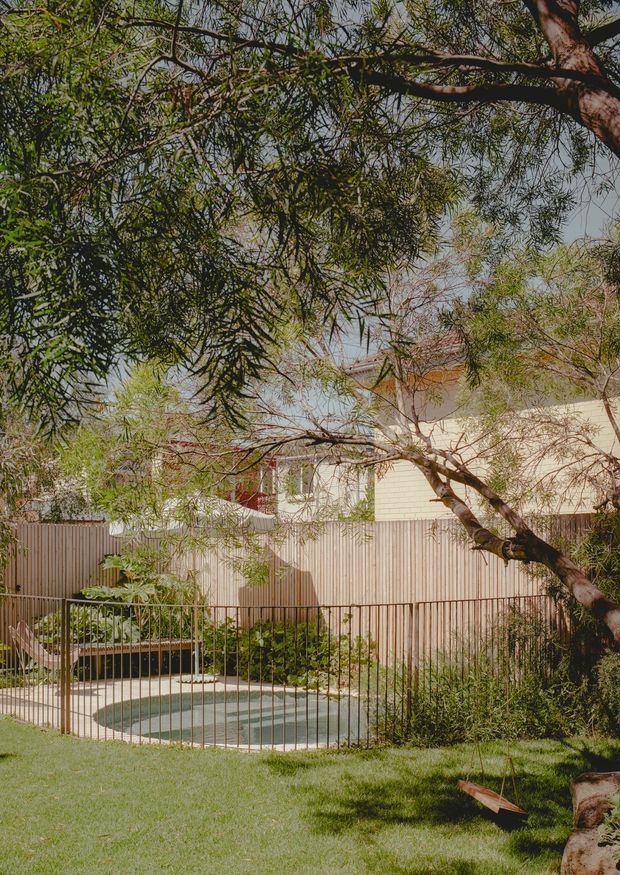 A landscaped residential backyard viewed through mature, leafy trees. The scene features a small rounded swimming pool enclosed by a dark metal fence, a timber-clad or weatherboard house with a pale facade and terracotta-toned roof glimpsed behind it, planted garden beds, lawn, and a timber garden swing. The image has a warm, natural, softly shaded atmosphere.