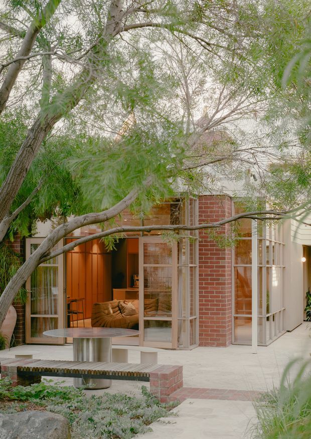 A tranquil courtyard view of a contemporary brick-and-glass residence framed by mature, feathery trees. The home features red brick walls, extensive white-framed glazing, warm timber or orange-toned interior finishes, and an open connection between the living area and paved outdoor courtyard. A built-in bench, round outdoor table, and landscaped planting occupy the foreground.