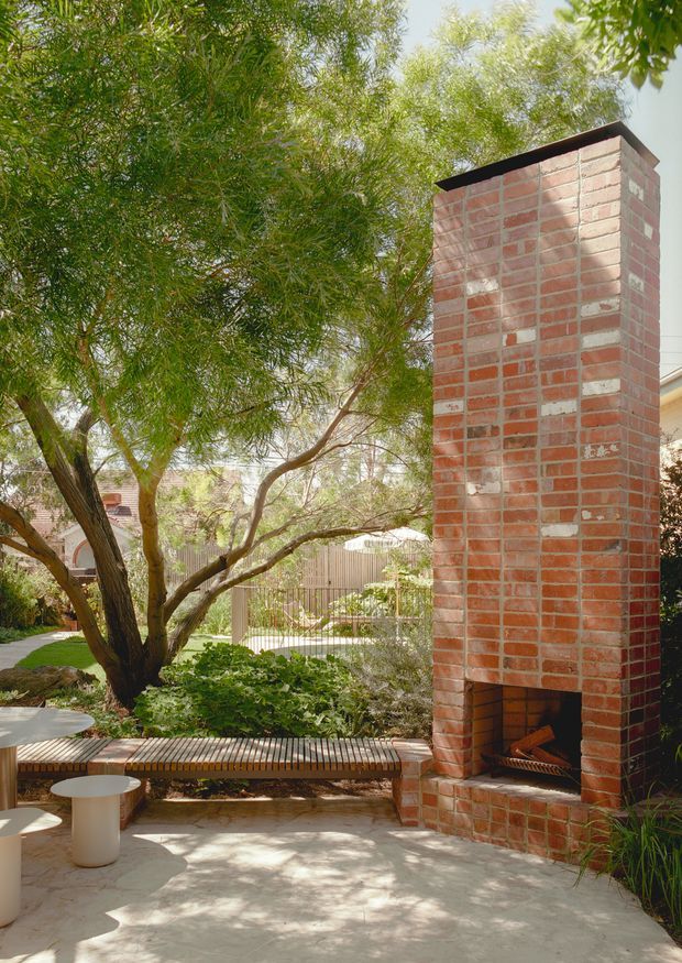 A sunlit landscaped courtyard or outdoor entertaining area featuring a tall exposed red brick fireplace with a dark metal cap and open firebox. A mature multi-trunk tree, dense planting, timber slatted built-in bench, pale paved flooring, and small round outdoor tables create a sheltered garden setting.