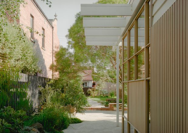 A landscaped residential passage beside a contemporary timber-clad building, with tall glazed windows, a projecting white pergola or canopy, and a paved path leading through a lush garden courtyard. A neighbouring brick building is visible on the left, while the foreground architecture and garden dominate the scene.