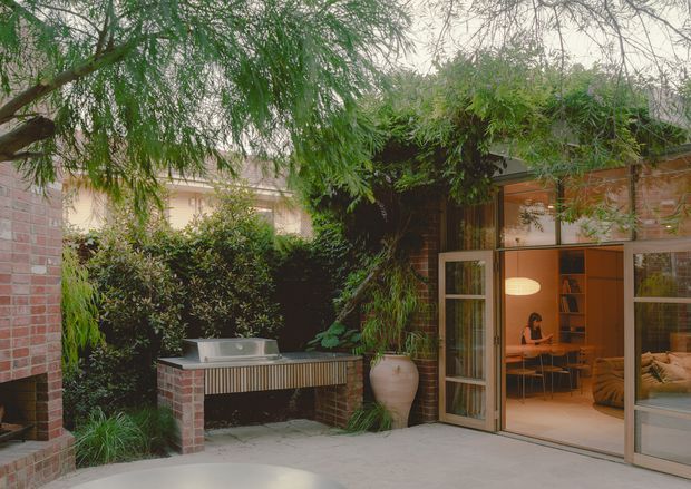 A leafy, sheltered courtyard viewed from outdoors, with a built-in brick barbecue or outdoor kitchen counter, lush planting, and large glazed timber-framed doors opening into a warm contemporary interior. The visible interior includes a dining area, shelving, pendant lighting, and a sofa.