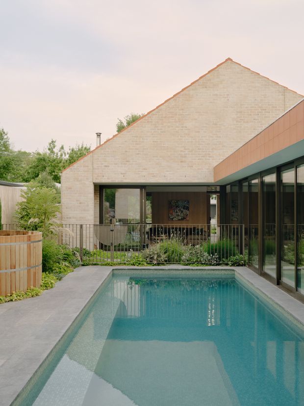 A contemporary residential courtyard viewed from beside a rectangular swimming pool. The house features pale exposed brick walls, a steep gabled roof, warm timber-clad fascia, extensive dark-framed glazing, and a covered outdoor seating area. Landscaped planting surrounds the pool and the architecture forms an enclosed, private courtyard.