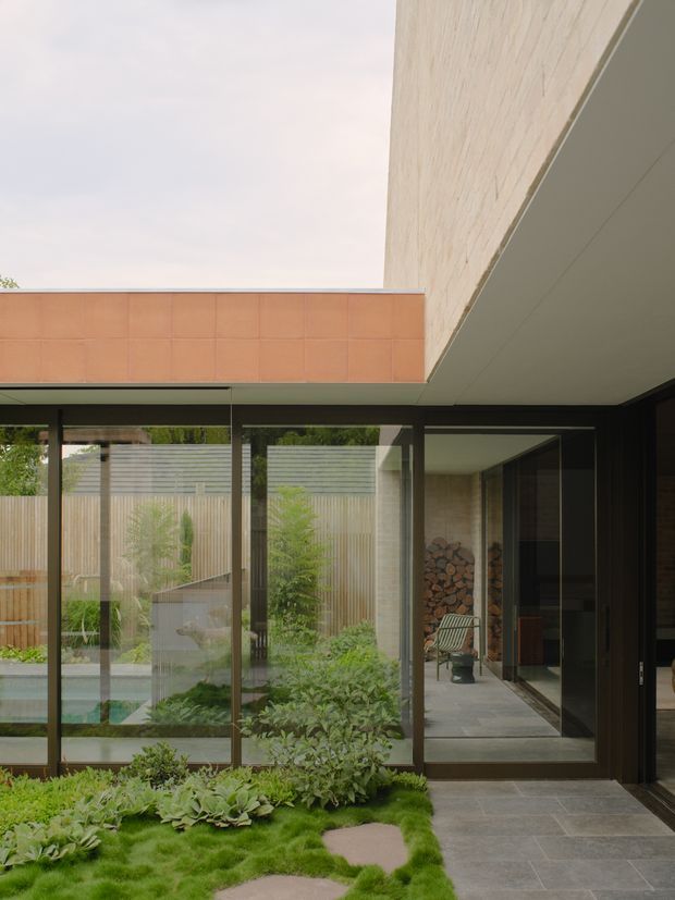 A contemporary courtyard viewed from beneath a covered architectural overhang, with expansive dark-framed glazing opening toward a planted garden and partially visible pool. The space features a tiled terrace, minimalist concrete or rendered surfaces, a warm terracotta tiled wall, timber screening, and an interior area with stacked firewood and a lounge chair. The composition emphasizes indoor-outdoor connection, clean horizontal and vertical lines, and a sheltered landscaped courtyard.