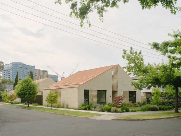 Street view of a low-rise modern building with a prominent pitched terracotta-tiled roof, pale textured masonry walls, dark window and door openings, and perforated breeze-block screens. The landscaped frontage includes established trees, shrubs, and a footpath, with taller urban buildings visible in the background.