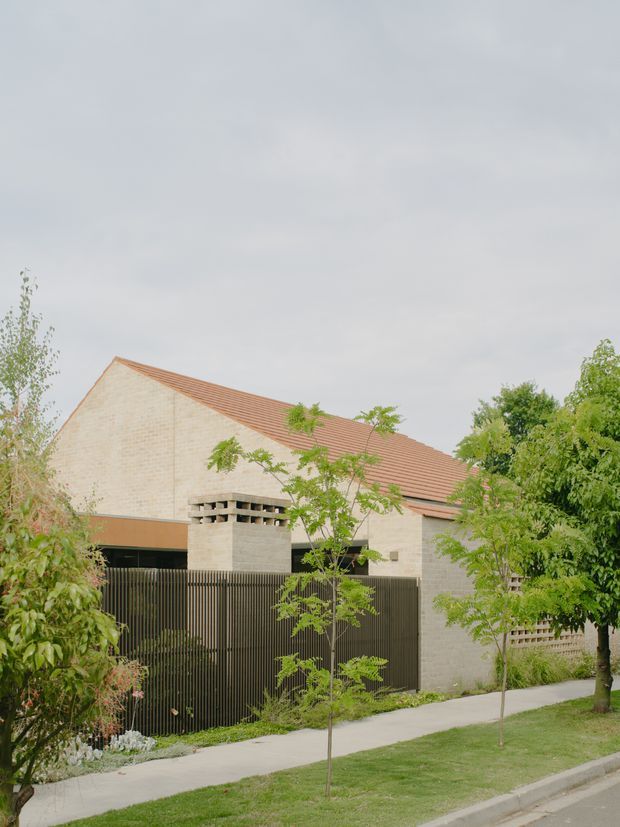 Exterior view of a low-rise residential building with a prominent pale brick gabled volume, terracotta tiled pitched roof, dark vertical slatted fencing and garage or entry opening. Young trees, landscaped planting and a concrete footpath line the street frontage.