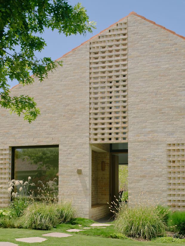 Exterior view of a contemporary single-storey brick building with a steep gabled roof and a prominent breeze-block screen above the recessed entry. Pale masonry walls, dark-framed glazing, and landscaped grasses surround the entrance, with a stepping-stone path across the lawn.