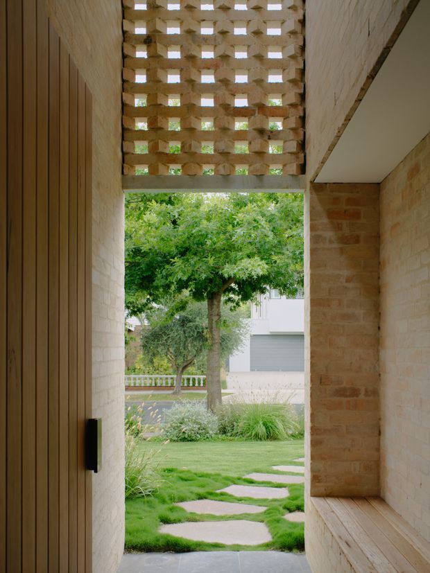 A narrow brick-lined entry passage opening onto a landscaped garden. The passage features a tall timber door on the left, a built-in timber bench on the right, and a perforated brick breeze-block screen overhead that filters daylight and views of the trees beyond. Stepping stones lead through the lawn outside.