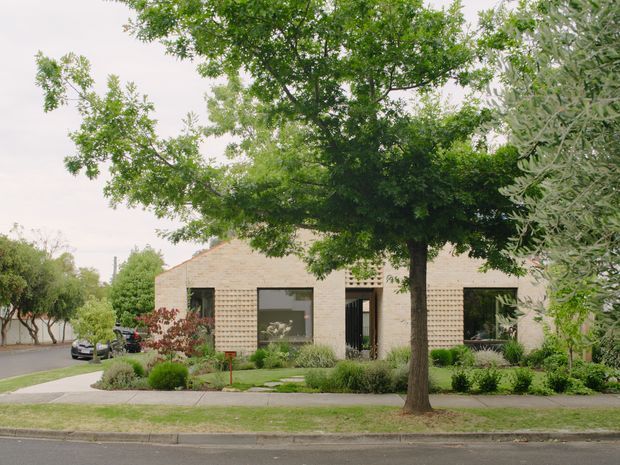 Street-facing view of a low-rise contemporary residential building set within a landscaped suburban garden. The facade uses pale cream brickwork with sections of patterned breeze-block screening, dark-framed windows and doors, and a simple low-pitched roofline. Mature deciduous trees and structured planting frame the property, with a footpath and quiet residential street in the foreground.