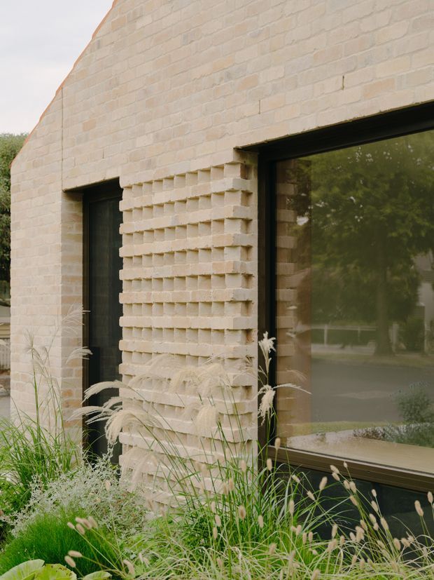 Close-up exterior view of a contemporary brick building facade with pale textured masonry, deeply recessed dark-framed openings, and a large reflective window. A projecting patterned brick section creates a tactile feature wall, partially softened by ornamental grasses and planting in the foreground.