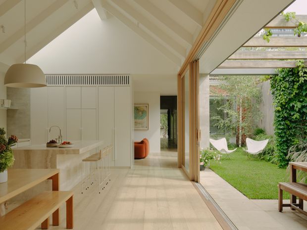 A bright, open-plan kitchen and living area with a high pitched, white-painted ceiling and exposed structural beams. The kitchen features white full-height cabinetry, a pale stone island and timber stools and dining bench. Large timber-framed sliding glass doors open directly onto a green courtyard with a pergola, climbing greenery and outdoor seating. The interior is light, warm and minimalist, with strong indoor-outdoor connection.