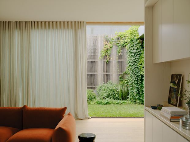 A softly lit contemporary living room with a rust-orange sofa, full-height cream curtains and pale timber flooring. The room opens directly to a lush private garden through a large glazed opening, while white built-in cabinetry and a countertop line the right wall. The garden includes a vertical slatted screen and climbing greenery.