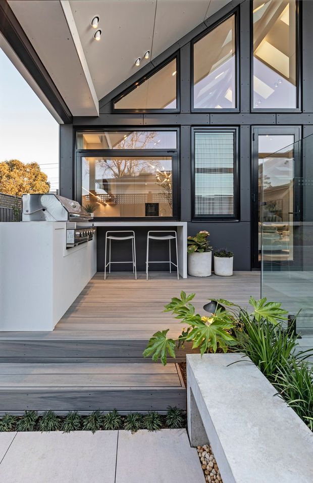 Contemporary outdoor entertaining area with a built-in barbecue and white benchtop, bar seating, timber decking, dark cladding, and extensive glazed doors and windows. The deck steps down toward a landscaped garden with concrete planters and ornamental planting.