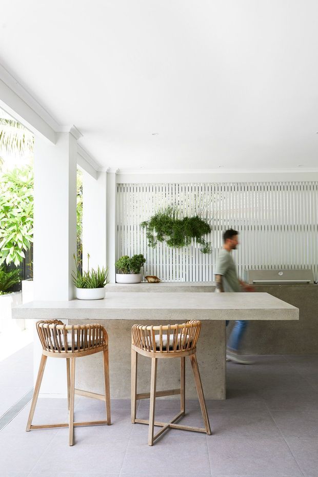 Bright, open-plan outdoor kitchen or entertaining area with a substantial concrete island and two timber bar stools. White structural columns and ceiling, grey tiled flooring, a vertical slatted privacy screen, integrated cabinetry, and abundant greenery create a clean contemporary setting. A person is blurred in the background near the counter.