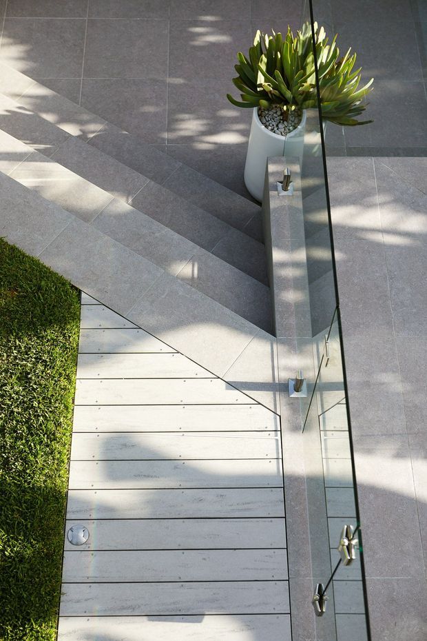 Top-down view of a contemporary outdoor transition area featuring large-format grey tiled paving, pale timber decking, a narrow strip of lawn, a frameless glass balustrade, and a tall white planter with architectural green foliage. Strong sunlight creates patterned shadows across the hardscape.